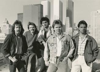 Five young men with shaggy hair in jeans on rooftop with downtown skyline behind them. Daytime,…