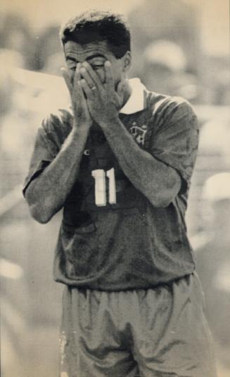 Man in soccer shorts and shirt has his hands over his face and eyes closed.