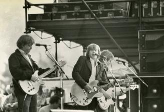 Three men with shaggy hair wearing dark jackets play guitars in front of microphones. The middl…