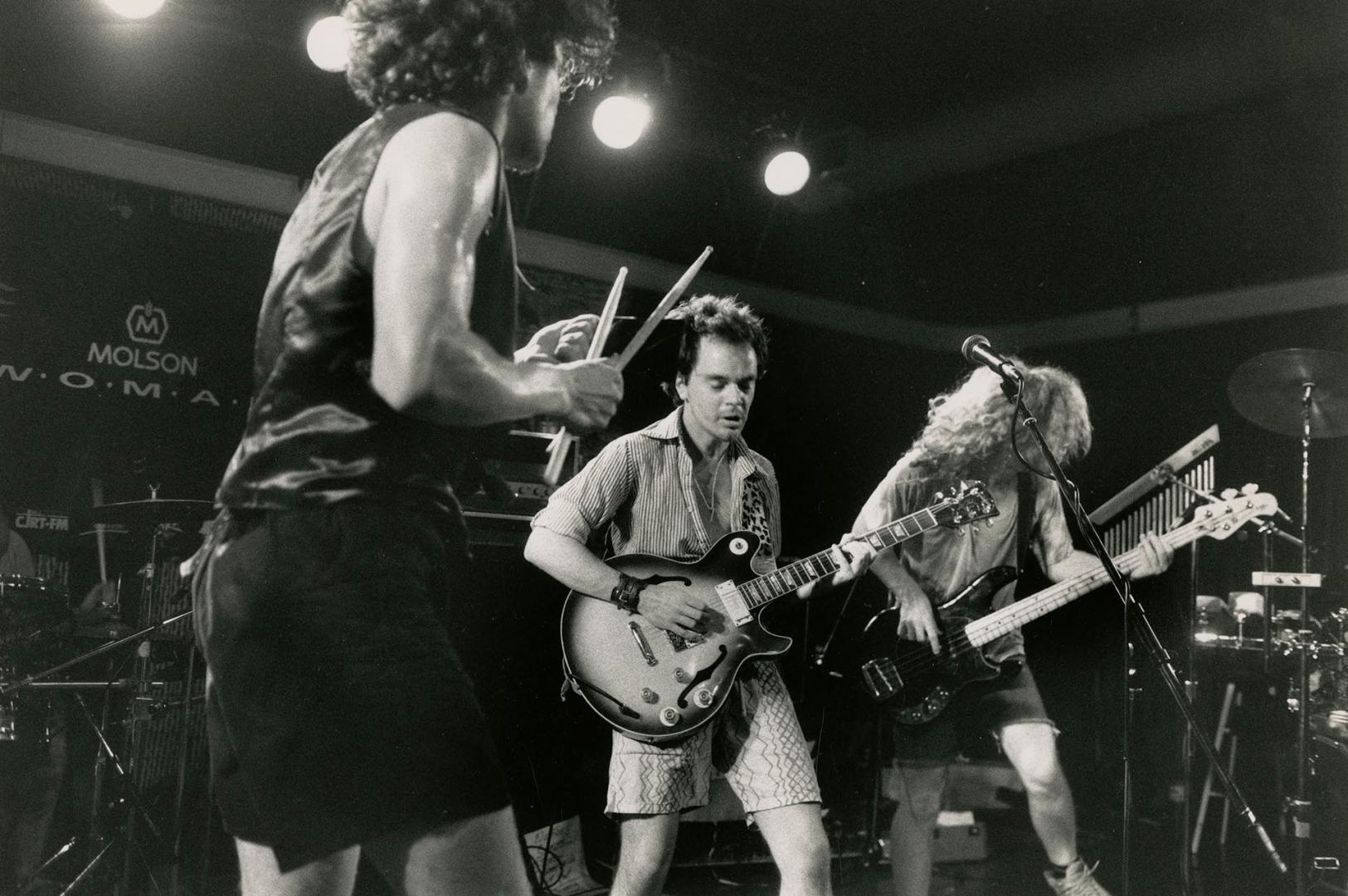 Three musicians on darkened stage. From left one has drumsticks on cowbell, next on electric gu…