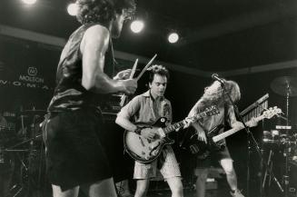 Three musicians on darkened stage. From left one has drumsticks on cowbell, next on electric gu…