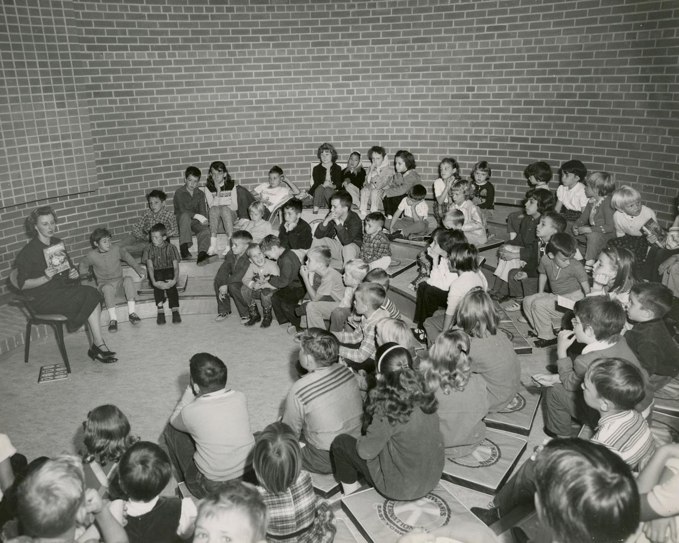 Picture of a librarian reading to a group of children seated in a circle around her. 