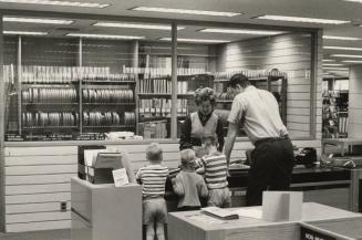 Picture of a librarian showing three children and a man something at a desk with a room full of…