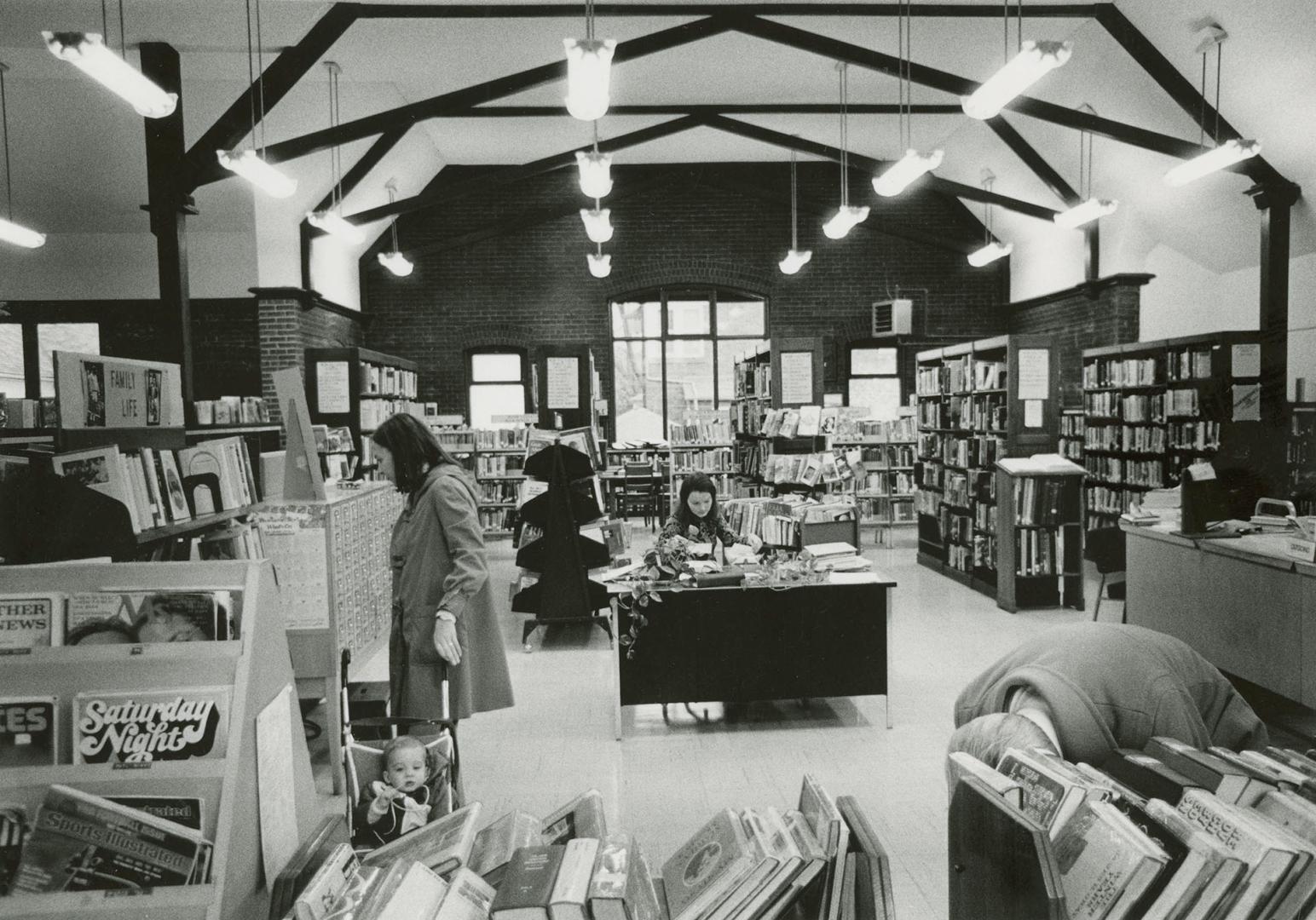 Picture of library interior with beamed ceiling and crowded bookshelves and woman with a child …