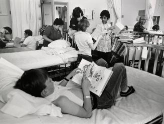 Picture of a large hospital room with a boy lying on his bed reading a book and several people …