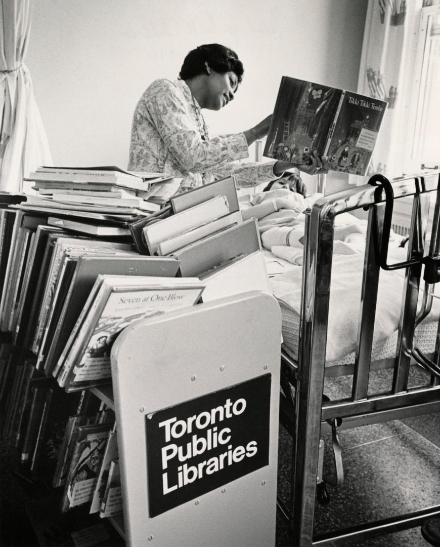 Picture of a librarian reading a book to a child lying in a hospital bed with full book truck i…