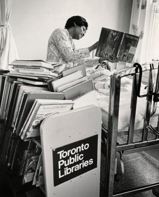 Picture of a librarian reading a book to a child lying in a hospital bed with full book truck i…