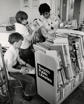 Picture of a librarian showing two boys books from an overflowing book truck. 