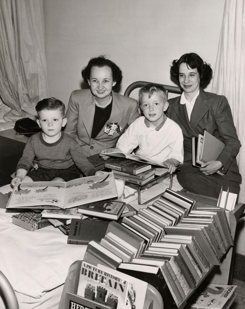 Picture of a two librarians sitting with two boys on a hospital bed looking at a pile of books …
