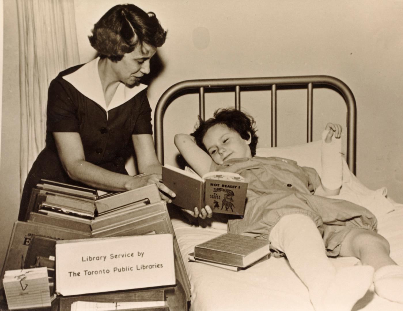 Picture of a librarian showing a book to a child in a cast lying in a hospital bed. 