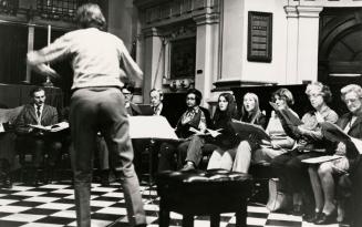 In an old ornate hall men and women sitting on pews hold sheet music and sing before a conducto…