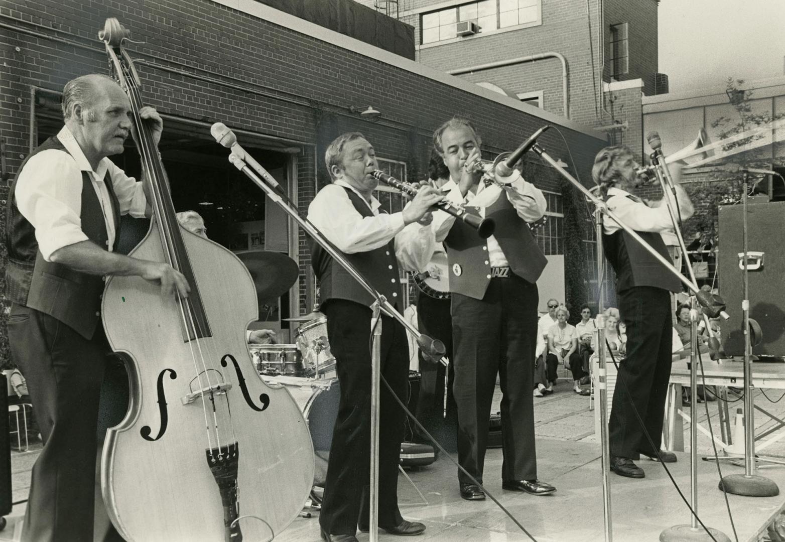 4 middle aged men in matching trousers and vests behind microphones on stands play front left t…