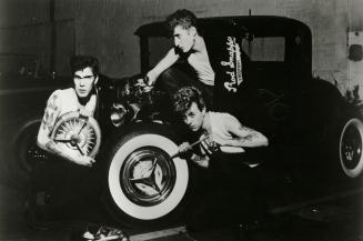 3 guys in white t-shirts hanging off an old hotrod car. 