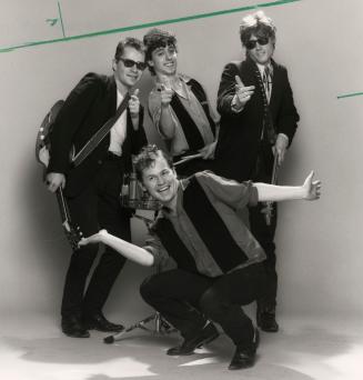 Four young men, one kneeling in front, wearing business casual clothing. Two carry guitars and …