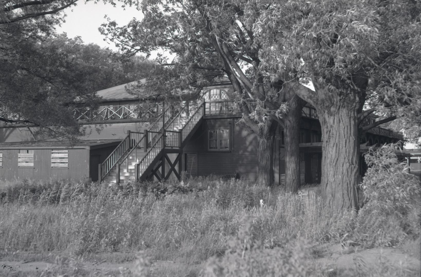 Photograph of an abandoned wooden dance pavilion with large trees in front and overgrown grasse…