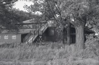 Photograph of an abandoned wooden dance pavilion with large trees in front and overgrown grasse…
