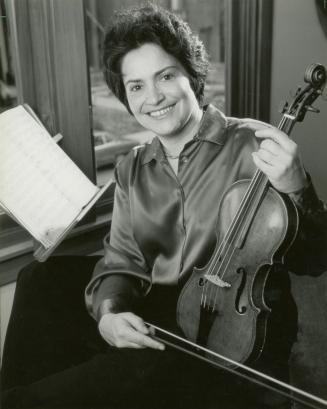 Smiling woman holding a violin and bow beside sheet music on stand. 