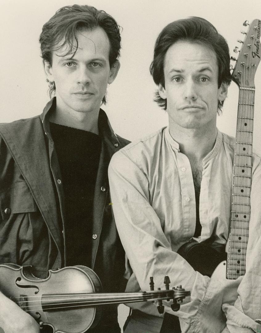 Two young men from torso, staring directly into the camera. On the left with violin in dark shi…