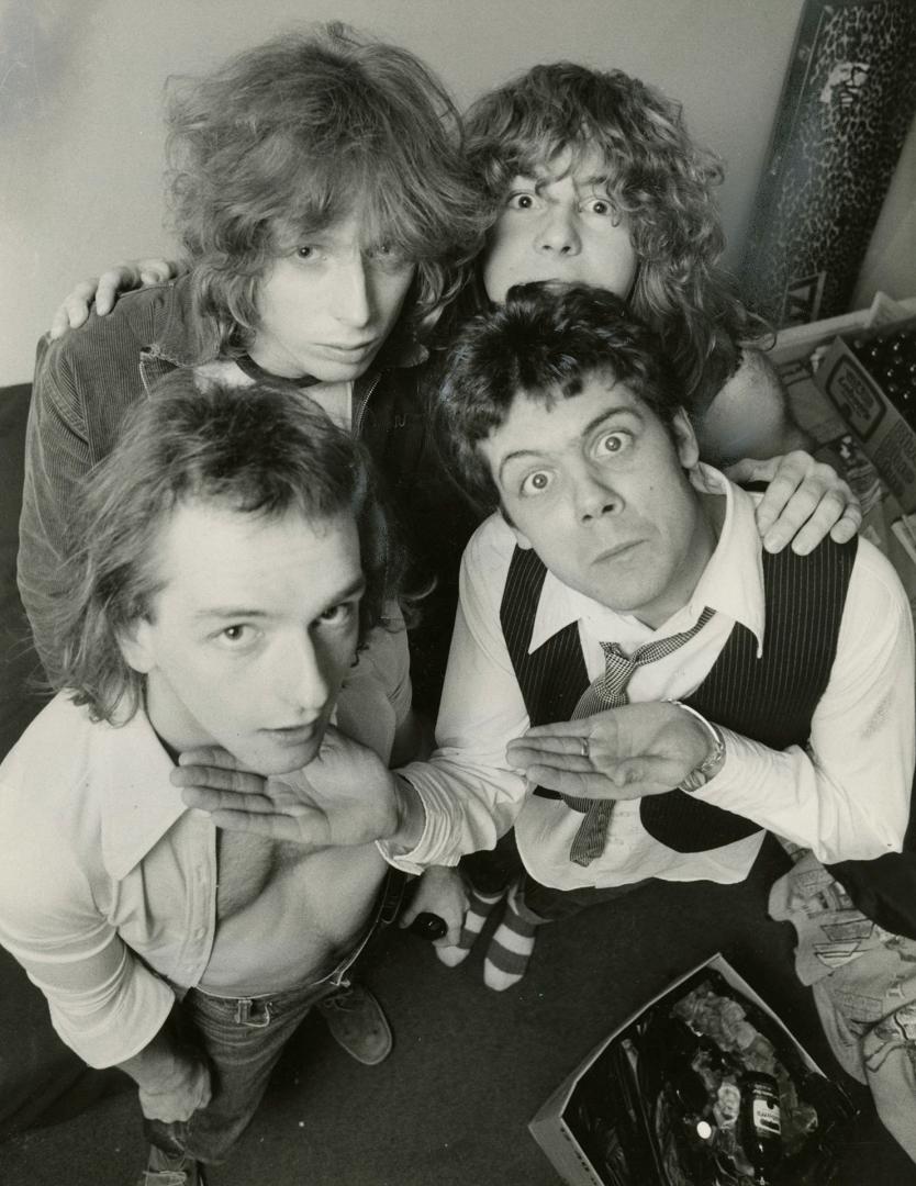 Four young men in a tight cluster look up into the camera, with shaggy hair and shirts on. 