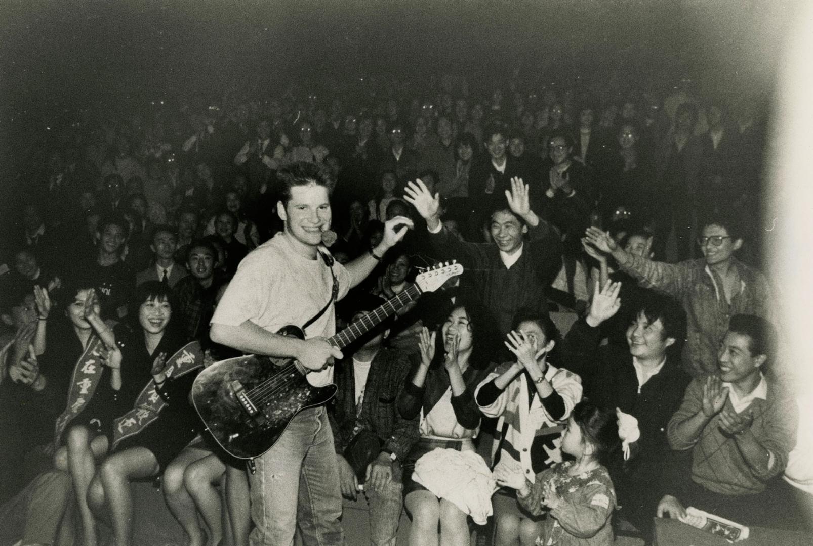 Clean cut young man with guitar looks back at camera with audience behind him. 