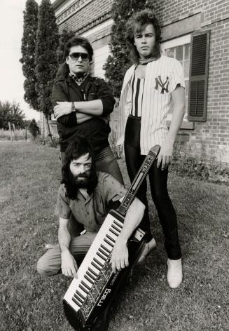 On grass in front of brick building, two men stand, one kneels while holding Moog keyboard. 