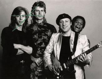 4 men, casually dressed pose against grey background. One in tam holds guitar.
