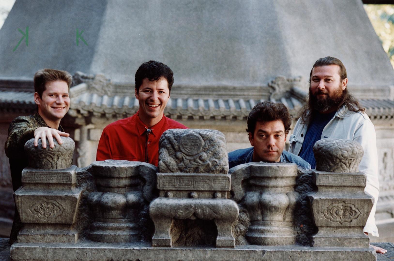 4 young men, casually dressed stand behind wall with Chinese building tiles.. Roof behind them.