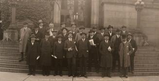 Black-and-white group photograph of about sixteen adults standing on stone steps outside a larg…