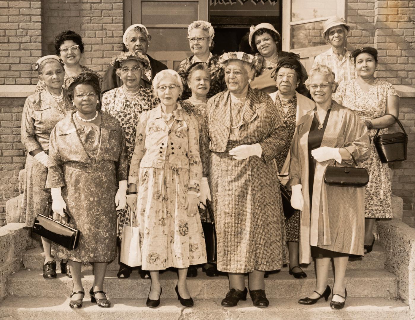 Black-and-white photograph of a group of fourteen women standing in three rows on exterior step…