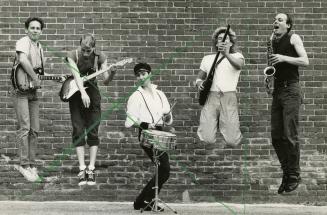 Outdoors, day, in front of brick wall, five men jump up in the air with their instruments excep…