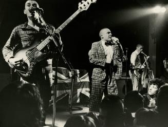 On dark stage, three young men with short hair dressed in plaid.. singer in middle, guitarists …