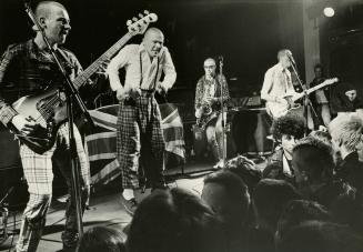 On dark stage, four young men with short hair dressed in plaid. The singer jumping up in middle…