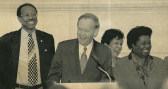 Against white wall, tall black man and shorter white with microphone man in suits, then two sho…